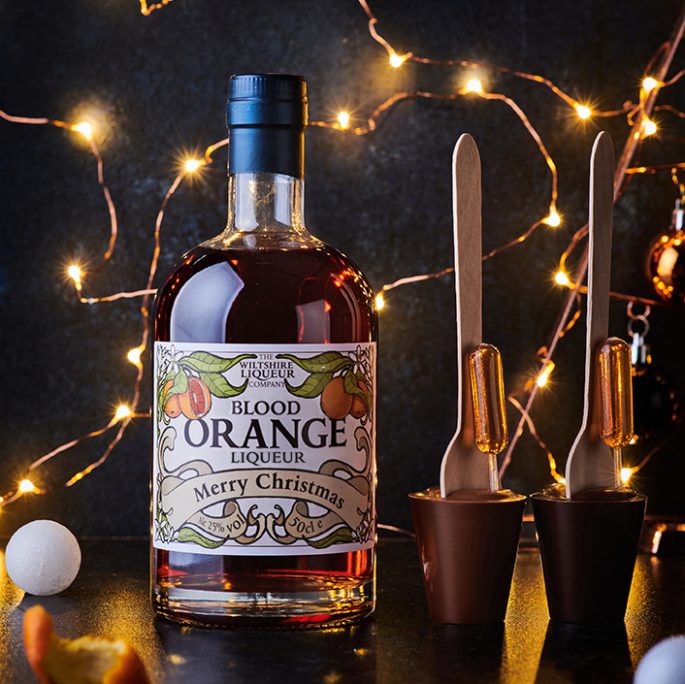 A Blood Orange Liqueur bottle and hot chocolate stirrers on a Christmasy background of fairy lights.