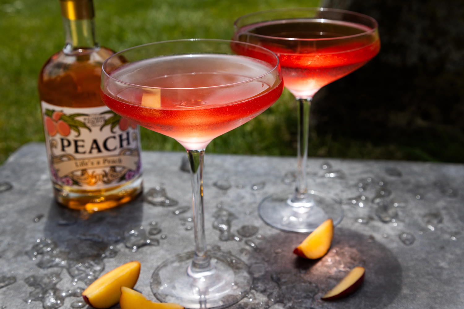Two martini glasses of Sanctuary cocktail surrounded by peach slices and made with The Wiltshire Liqueur Company's Peach Liqueur.