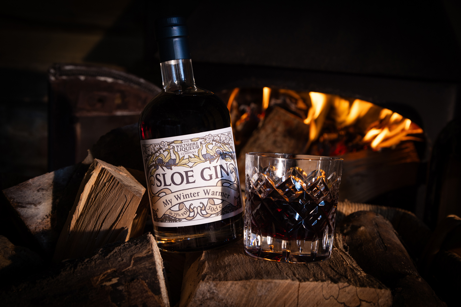 A personalised bottle of Sloe Gin that reads, 'my winter warmer' sits by an open fire. A glass of Wiltshire Liqueur's Sloe Gin sits close by.