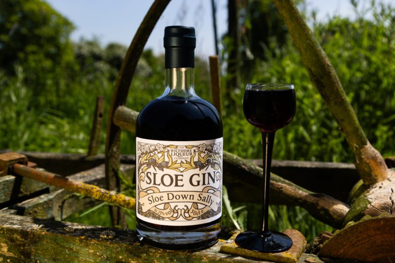 A Bottle of The Wiltshire Liqueur Company's Sloe Gin sits on a rustic wheel in the countryside. The personalised label reads: 'Sloe Down Sally'.